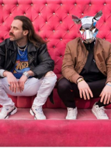 Dwóch mężczyzn kuca, za nimi różowa ściana. Jeden z nich ma na głowie srebrną maskę szczura.