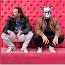 Dwóch mężczyzn kuca, za nimi różowa ściana. Jeden z nich ma na głowie srebrną maskę szczura.