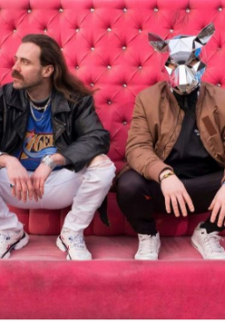 Dwóch mężczyzn kuca, za nimi różowa ściana. Jeden z nich ma na głowie srebrną maskę szczura.