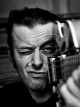 Tymon Tymański trzyma gitarę tak, jakby to była broń i celuje nią przed siebie mrużąc jedno oko