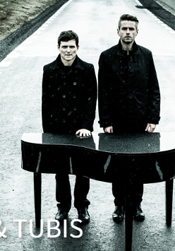 Dwóch mężczyzn ubranych na czarno, stoją na ulicy za małym fortepianem.