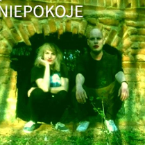 Kobieta i mężczyzna kucają przy murze. Mają pomalowane na czerwono usta.