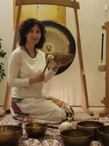 Kobieta w białej bluzce siedzi na dywanie. Przed nią stoją misy i gongi do grania.