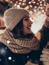 Kobieta i mężczyzna w zimowych straojach uśmiechają się do siebie, a w ręku mają kubki na gorące napoje.