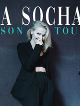 Uśmiechająca się kobieta zasłania lekko twarz ręką. Jest ubrana w czarny garnitur i stoi na tle czarnych metalicznych plandek.