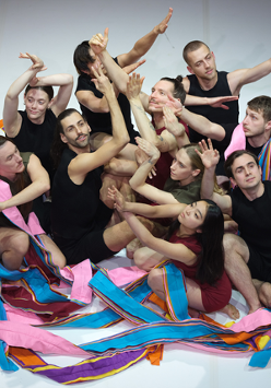 Zdjęcie grupy tancerzy siedzących na podłodze, z uniesionymi rękoma; niektórych z nich oplatają kolorowe szarfy.