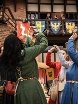 Grupa osób w średniowiecznych strojach tańczy na tle zamku i herbów.