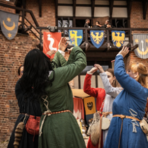 Grupa osób w średniowiecznych strojach tańczy na tle zamku i herbów.