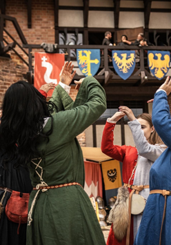 Grupa osób w średniowiecznych strojach tańczy na tle zamku i herbów.