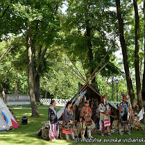 Indianie w charakterystycznych dla ich kultury strojach oraz namioty tipi. W tle trawa, drzewa i biały płot.