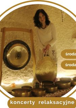 Kobieta w średnim wieku, w białym stroju, z czarnymi kręconymi włosami. Stoi przy rozłozonych złotych misach, trzyma duży gong w ręce. W tle wisi drugi gong.