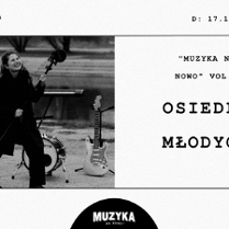 Czarno-biała fotografia przedstawia kobietę w długim płaszczu, która się uśmiecha i trzyma mikrofon na statywie. Za nią znajdują się różne instrumenty muzyczne: gitara basowa, gitara akustyczna, perkusja i gitara elektryczna. W tle woda i schody.