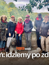 Grupa ludzi pozująca do zdjęcia z kilkoma psami. W tle drzewa oraz jezioro. Na froncie tytuł wydarzenia "Power walk" i hasło: "Poznań: idziemy po zdrowie!"
