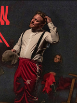 Zdjęcie przedstawia scenę teatralną w ciemnej tonacji. Na pierwszym planie stoi mężczyzna w białej koszuli z szelkami i czerwonych spodniach dresowych, trzymający w dłoni kaszkiet i drapiący się po głowie. W tle, w półmroku, widoczne są dwie inne postacie oraz fragment drewnianego stołu. W górnym lewym roku tytuł spektaklu zapisany czerwoną, niejako rozmytą czcionką.