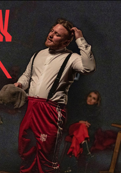 Zdjęcie przedstawia scenę teatralną w ciemnej tonacji. Na pierwszym planie stoi mężczyzna w białej koszuli z szelkami i czerwonych spodniach dresowych, trzymający w dłoni kaszkiet i drapiący się po głowie. W tle, w półmroku, widoczne są dwie inne postacie oraz fragment drewnianego stołu. W górnym lewym roku tytuł spektaklu zapisany czerwoną, niejako rozmytą czcionką.