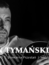 Czarno-białe, bliskie ujęcie mężczyzny z krótkim zarostem, który patrzy w bok z lekkim uśmiechem. W dolnej części kadru widoczny jest gryf i fragment korpusu gitary akustycznej trzymanej przez artystę. Tło jest ciemne i nieostre. Na froncie napisy, będące szczegółami wydarzenia, które są zawarte w artykule.