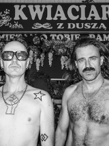 Czarno-białe zdjęcie przedstawia dwóch mężczyzn stojących przed kwiaciarnią z szyldem "Kwiaciarnia z duszą". Mężczyzna po lewej ma ogoloną głowę, ciemne okulary, tatuaże na klatce piersiowej i ramieniu. Mężczyzna po prawej ma długie włosy i wąsy. Obaj są bez koszulek.