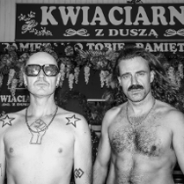 Czarno-białe zdjęcie przedstawia dwóch mężczyzn stojących przed kwiaciarnią z szyldem "Kwiaciarnia z duszą". Mężczyzna po lewej ma ogoloną głowę, ciemne okulary, tatuaże na klatce piersiowej i ramieniu. Mężczyzna po prawej ma długie włosy i wąsy. Obaj są bez koszulek.