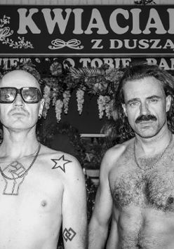 Czarno-białe zdjęcie przedstawia dwóch mężczyzn stojących przed kwiaciarnią z szyldem "Kwiaciarnia z duszą". Mężczyzna po lewej ma ogoloną głowę, ciemne okulary, tatuaże na klatce piersiowej i ramieniu. Mężczyzna po prawej ma długie włosy i wąsy. Obaj są bez koszulek.