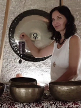 Kobieta w średnim wieku, w białej koszule, z czarnymi kręconymi włosami. Stoi przy rozłozonych złotych misach.