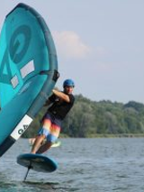 Zdjęcie osoby uprawiającej wingfoiling na jeziorze. Sportowiec, ubrany w kask i kolorowe spodenki, unosi się nad powierzchnią wody na desce. W dłoniach trzyma duży, turkusowy żagiel z ciemnymi detalami, który jest wygięty pod wpływem wiatru. W tle widać gęstą linię zielonych drzew na brzegu oraz jasne, pogodne niebo.