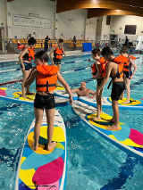 Uczestnicy zajęć stoją w kapokach na deskach SUP. Trener w wodzie.