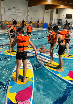 Uczestnicy zajęć stoją w kapokach na deskach SUP. Trener w wodzie.