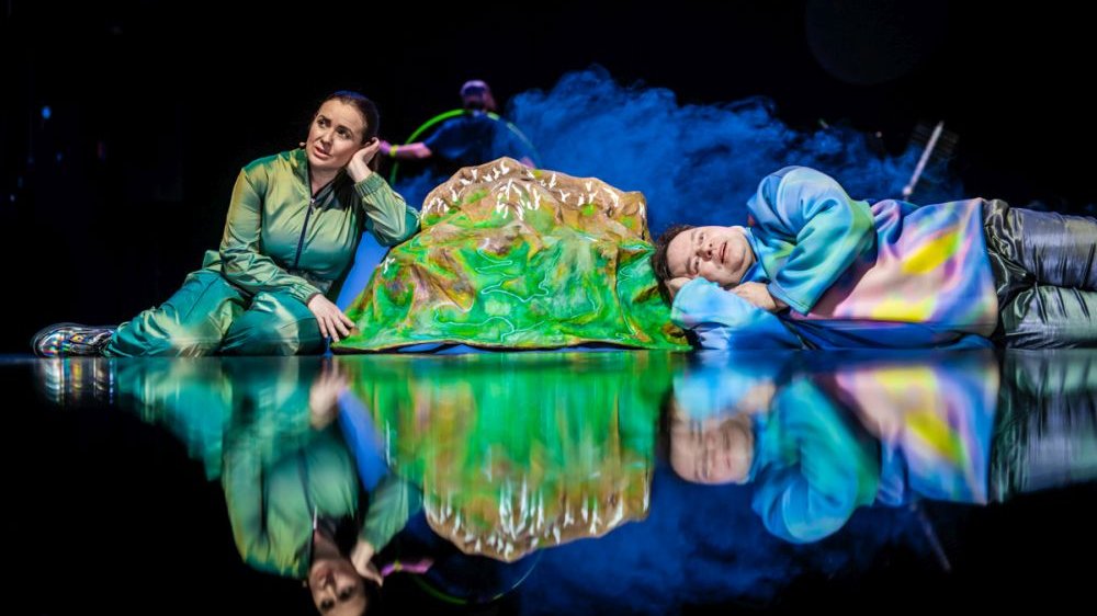 Dwoje artystów leży na scenie, opierając się o zieloną masę na środku. Mają na sobie błyszczące, rozświetlone na kolorowo ubrania, za nimi snuje się dym.