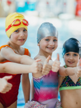 Grupa dzieci w strojach pływackich i czepkach stoi na basenie.