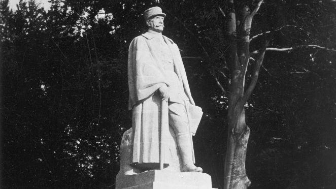 Czarno-białe zdjęcie pomnika. Na cokole pomnikowa postać mężczyzny w długim płaszczu i kapeluszu. U dołu stoi dwóch mężczyzn, patrzą w górę.