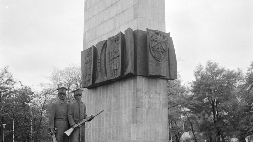 Czarno-biała fotografia pomnika. Czterokątna kamienna bryła z motywem żołnierzy i godła, po lewej kamienne figury dwóch uzbrojonych żołnierzy.