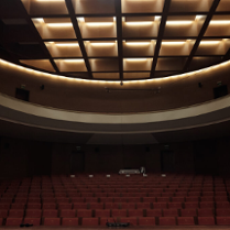 Obrazek przedstawia ciemną aule z czerwonymi krzesłami