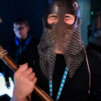 Osoba w wikińskim hełmie trzyma w obu dłoniach lekki toporek bojowy.