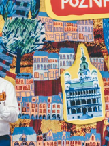 Uśmiechnięty mężczyzna gra na ukulele przed kolorową planszą, na której widać rysunki różnych znanych miejsc w Poznaniu.