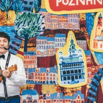 Uśmiechnięty mężczyzna gra na ukulele przed kolorową planszą, na której widać rysunki różnych znanych miejsc w Poznaniu.