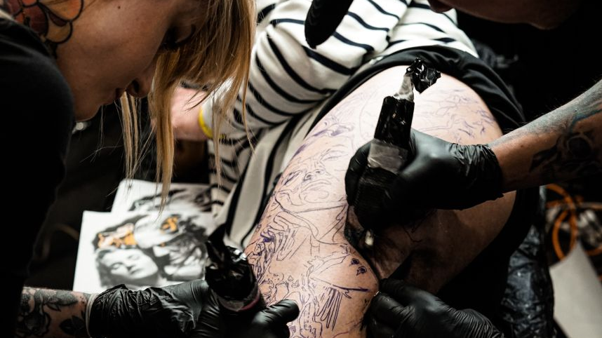 Dwoje tatuażystów tatuuje nogę leżącego chłopaka.