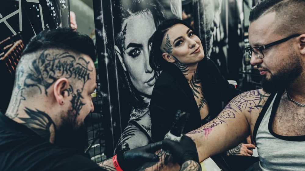 Dwóch mężczyzn i jedna kobieta podczas pracy tatuażysty. Kobieta uśmiecha się do zdjęcia, mężczyźni są zajęci tatuowaniem.