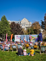 Ludzie siedzą na zielonej trawie w parku. Patrzą na scenę, na której występują ludzie. Scena jest ozdobiona logo, kolorowymi rysunkami.
