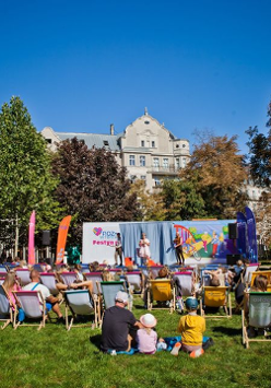 Ludzie siedzą na zielonej trawie w parku. Patrzą na scenę, na której występują ludzie. Scena jest ozdobiona logo, kolorowymi rysunkami.