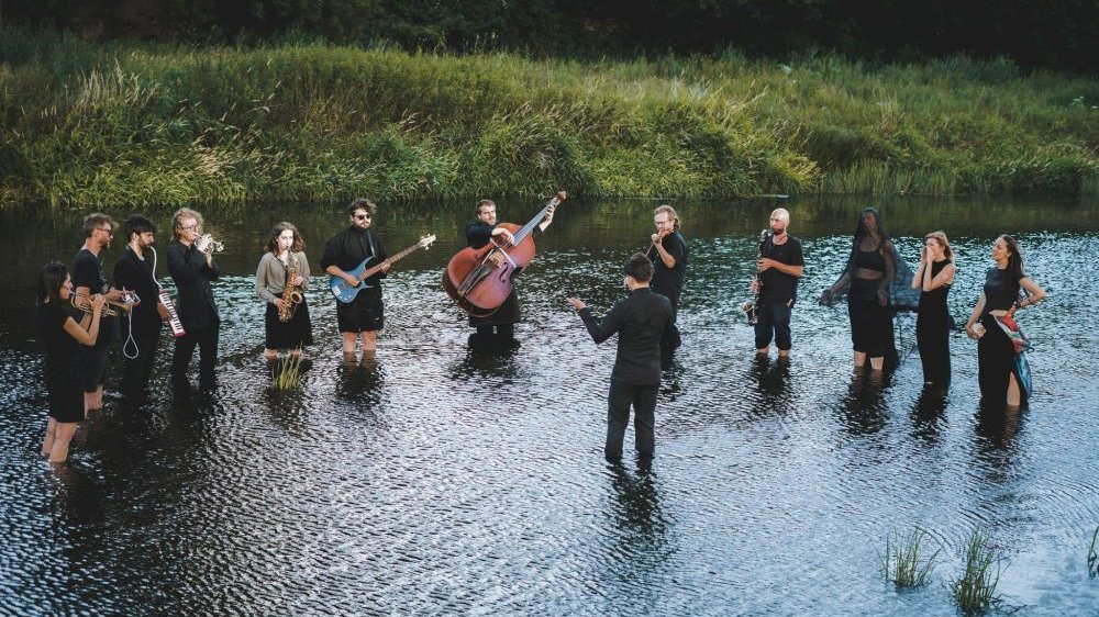 Kilkunastu członków orkiestry stoi w wodzie trzymając w rękach instrumenty muzyczne.