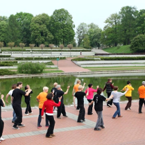 Osoby w różnym wieku stoją nad wybrzeżem stawu na Cytadeli i ćwiczą ruchy Tai Chi. W tle drzewa parku i reszta ścieżki nad stawem.