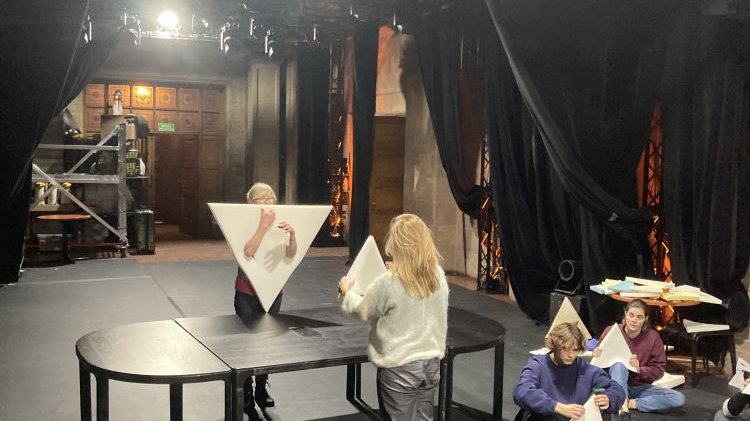Aktorzy i reżyserka na scenie. Dwie kobiety stoją naprzeciwko siebie nad czarnym stołem, w rękach trzymają wykonane z pianki trójkąty. Reszta siedzi na podłodze.