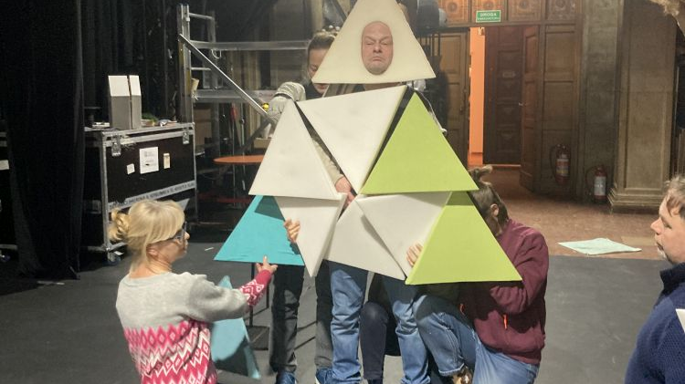 Aktorzy na próbie. Na środku stoi mężczyzna otoczony piankowymi trójkątami, przypomina bożonarodzeniową choinkę.