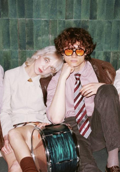 Kilka mlodych osób siedzi oparta o ścianę, oświetlona mocnym, białym światłem.