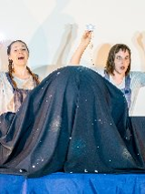 Obraz przedstawia dwie kobiety z emocjami na twarzach. Ich ręce są uniesione do góry. Przed nimi znajduje się trójwymiarowy obiekt pokryty tkaniną z gwiazdami.