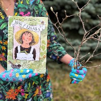 Ogrodniczka w jednej ręce trzyma książkę a w drugiej gałązki.