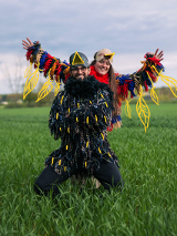 Kobieta i mężczyzna przebrani za kolorowe ptaki pozują do zdęcia. Stoją na zielonej polanie, za nimi widok na las.