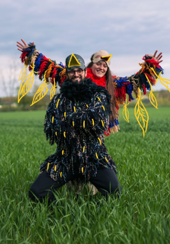 Kobieta i mężczyzna przebrani za kolorowe ptaki pozują do zdęcia. Stoją na zielonej polanie, za nimi widok na las.