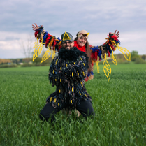 Kobieta i mężczyzna przebrani za kolorowe ptaki pozują do zdęcia. Stoją na zielonej polanie, za nimi widok na las.