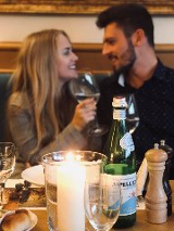 Zdjęcie, para siedzi przy zastawionym stole, świeca, kwiaty, butelka wina.